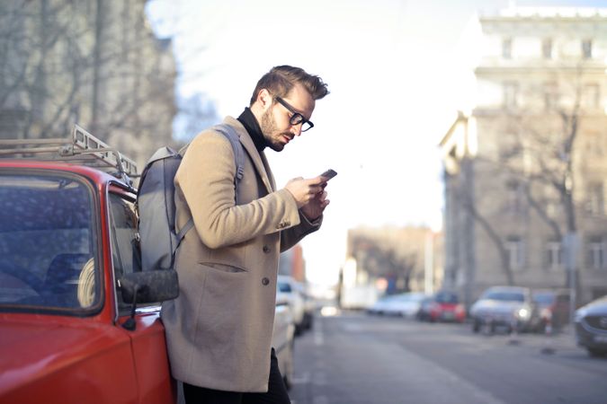 Man In Beige Coat Holding Phone Leaning On Red Vehicle Photo