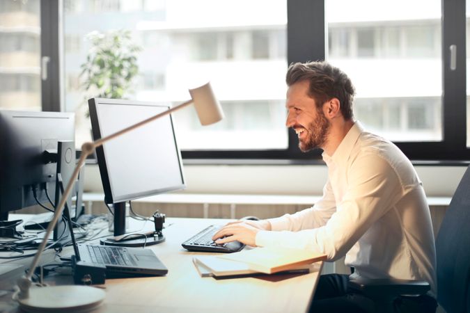 Man In White Dress Shirt Sitting On Black Rolling Chair While Facing Black Computer Set And Smiling Photo