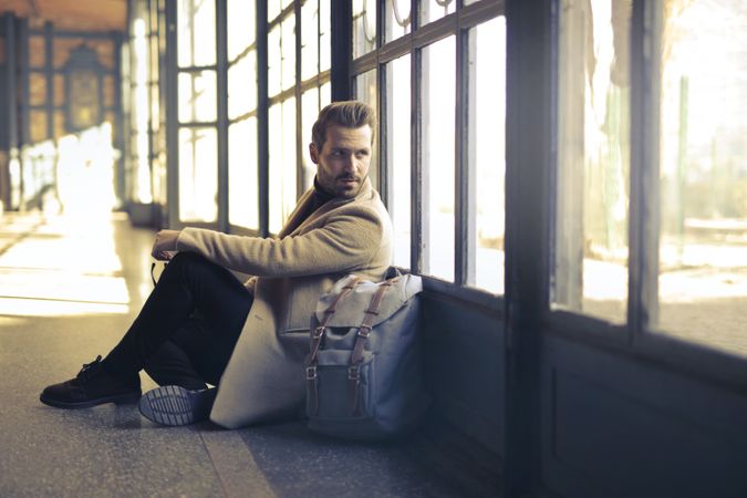 Man Wearing Brown Coat Beside White Backpack Sitting Near Window Photo