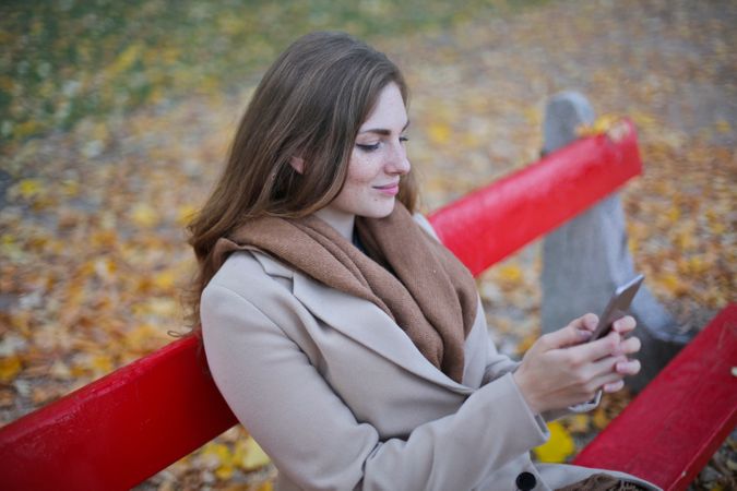 Woman In Beige Coat Holding Smartphone Sitting On Bench Photo