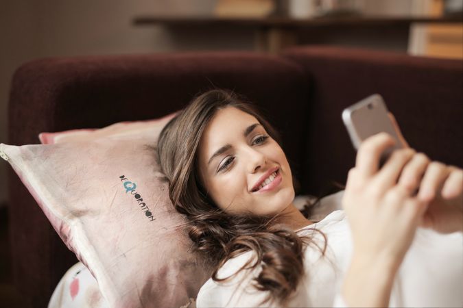 Woman In White Top Holding Smartphone Lying On Couch Photo