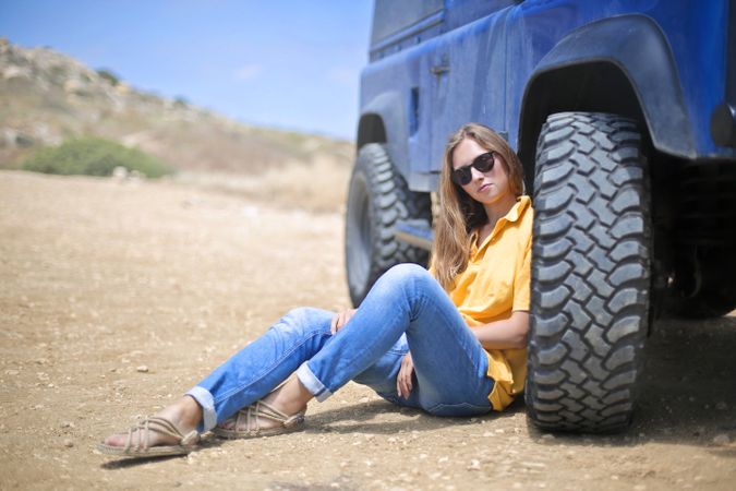 Woman In Yellow Polo Shirt Sitting On Ground Leaning On Blue Vehicle At Daytime Photo