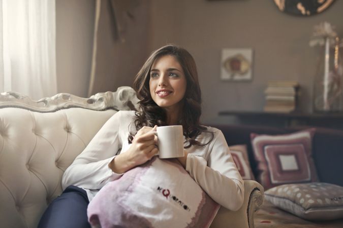 Woman Wearing White Top Drinking Beverage From White Ceramic Mug While Lying On Sofa Inside Well Lit Room Photo