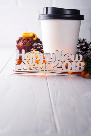 Paper Cup Of Coffee With Muffin, Pine Cone And Mandarin Surrounded By Lettering Happy New Year On White Wooden Background Photo
